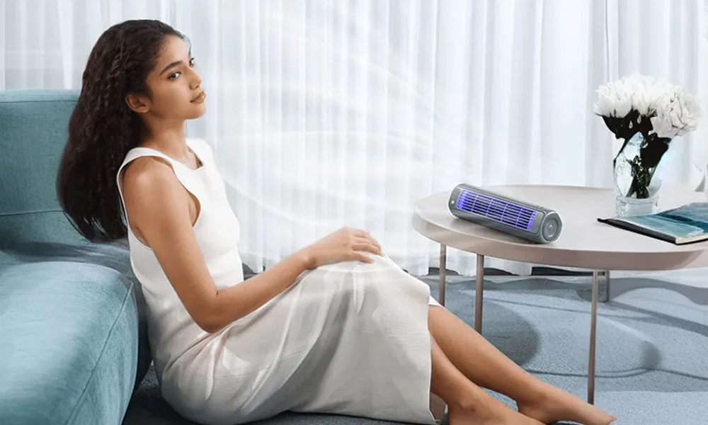 Elegant woman in a flowing white dress relaxing on a blue sofa in a bright, modern living room with sheer curtains, the nexo7 portable cooling device on the side table gently circulating cool air